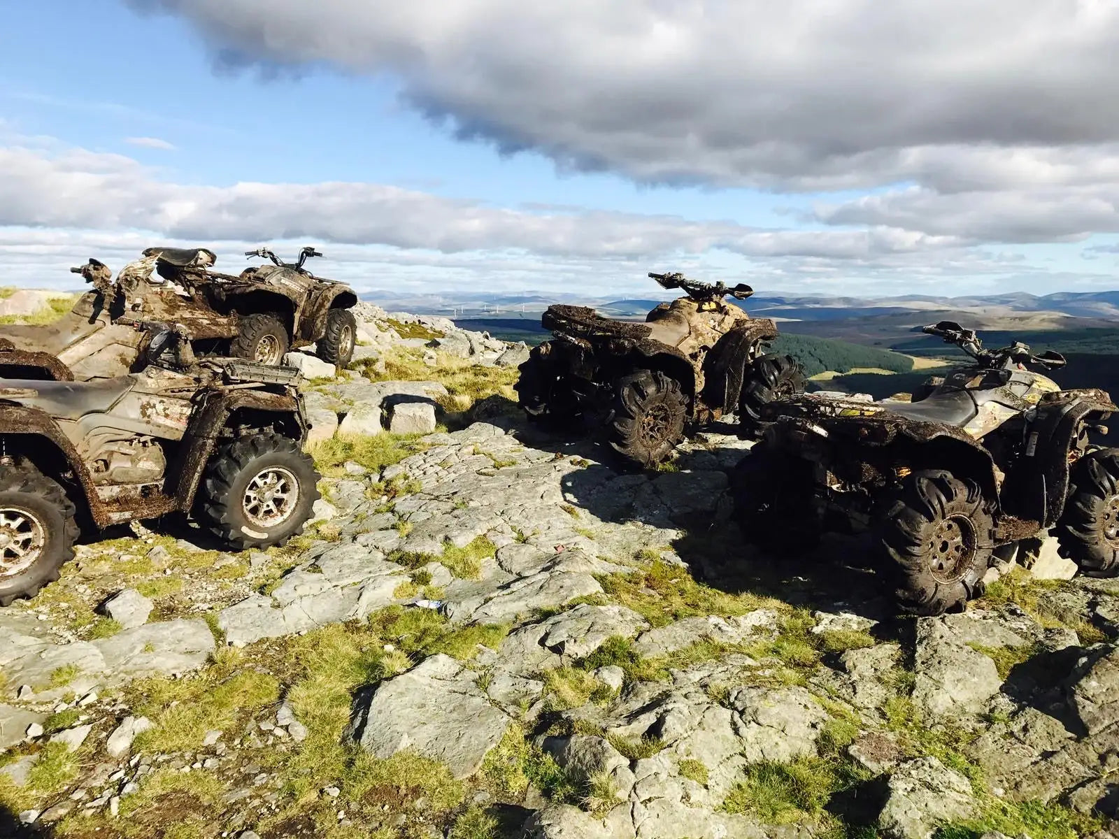 Three muddy, rugged all-terrain vehicles parked on a rocky hilltop under a partly cloudy sky.